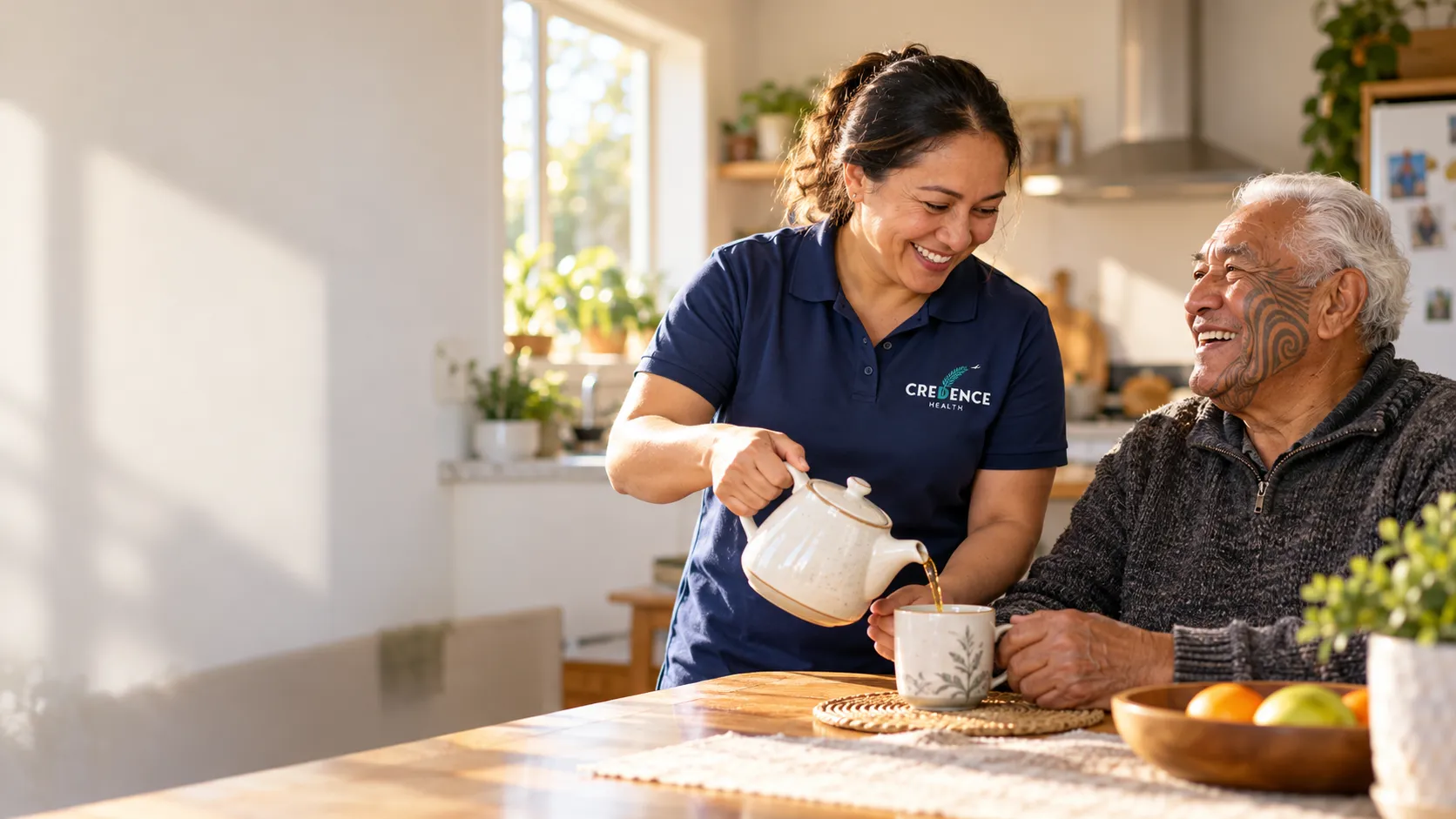 A home care worker in a navy Credence Health top pours tea for an older Māori man at his kitchen table